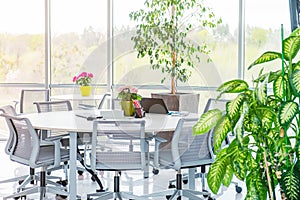 Round table as work place with laptop, notebooks in modern light open space office with big windows, desks, chairs and green plant