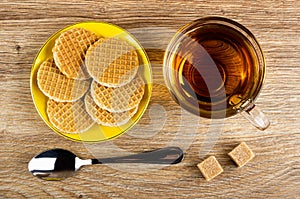 Round wafers with filling in saucer, sugar, spoon, cup of tea on table. Top view