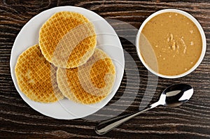 Round wafers in plate, bowl with peanut paste, spoon on wooden table. Top view