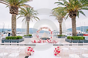 Round wedding arch stands on a pier with palm trees in front of rows of white chairs