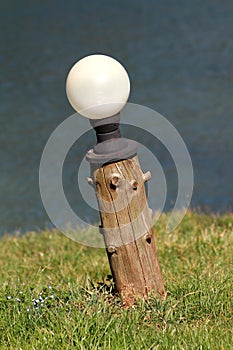 Round white backyard outdoor lamp mounted on tree stump leaning to one side surrounded with uncut grass and river in background