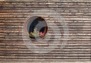 Round window with a geranium flower pot on  rustic wooden facade.