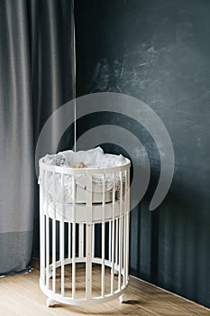 Round wooden bed for babies in the interior of the room against the background of a dark chalky wall. Convenient modern transforme