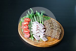 On a round wooden Board is sliced black bread with seeds, smoked brisket, green onions, lettuce, tomatoes and dill. Black photo