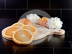 Round wooden cutting board with a handle with a fresh orange sliced up and arranged on it with 3 artificial white and orange