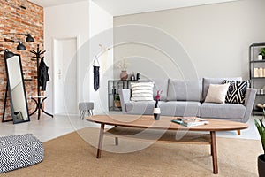 Round wooden table in the middle of elegant living room with grey sofa, metal shelf and mirror, real photo with copy space