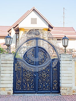 Rounded iron gates with a gilded wrought pattern and a fence enclosing a cottage