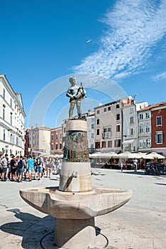 ROVINJ, CROATIA-AUGUST 30, 2018: Tourists walk by the Fountain with a sculpture of a boy and a fish and a clock Tower on