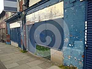 A row of abandoned stores with boarded up shop fronts with crumbling facades and peeling blue paint in an urban street