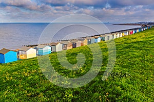 Row of beach huts along the coast in Tankerton, Whitstable, Kent. The green grass slopes are behind the huts and groynes