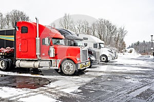 Row of big rigs industrial semi trucks with semi trailers standing on the winter truck stop parking lot with snow and ice