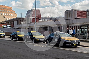 Row of black and yellow taxi cabs waiting for customers on taxi rank outside terminal building