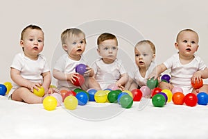 Row of caucasian babies sitting side by side looking away isolated on gray background. Five cute babies playing with