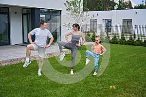 Row of caucasian mother, father and daughter doing leg exercise on green yard