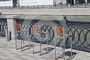 A row of chairs with wooden seats covered with snow near a metal fence on the canal embankment.