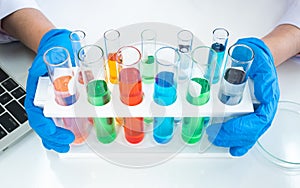 Row of colorful liquid chemical in test tube with scientist hand holding while wearing rubber blue gloves with laptop keyboard and
