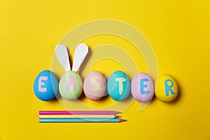 Row of colorful pastel monophonic painted Easter eggs with inscription Easter, fun bunny ears on egg, three pencils on