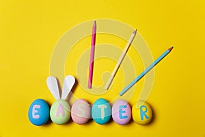 Row of colorful pastel monophonic painted Easter eggs with inscription Easter, fun bunny ears on egg, three pencils on