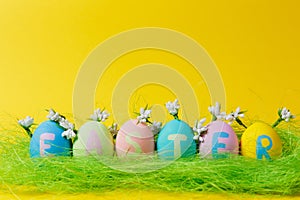 Row of colorful pastel painted Easter eggs with inscription Easter in green grass, white flowers lilies of valley