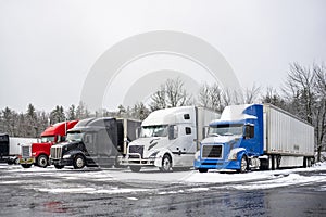 Row of the different big rigs semi trucks with loaded semi trailers standing on the winter truck stop parking lot with snow and