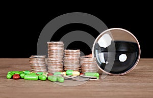 A row of dollar coins, a magnifying glass and capsule medicines scattered on the table in front of a dark background