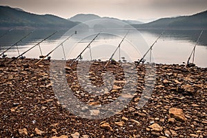 Row of fishing rods on a stony beach with mountains and lake on the background on a misty morning.