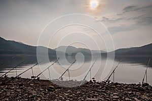 Row of fishing rods on a stony beach with mountains and lake on the background on a misty morning during sunrise.