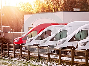 Row one red and many white commercial vans in a dealership for sale or rent in a snow. Cold winter season. warm sun flare. Used