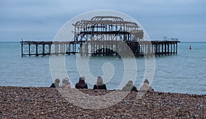 Row of people sitting on pebbly beach in Brighton UK on a wintry afternoon in December, in front of the ruins of West Pier.