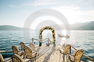 Row of soft chairs in front of a wedding arch on a cobbled pier with the sea and mountains in the background