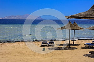 A row of straw umbrellas to protect against overheating and sunbeds on a sandy beach against a blue sky and blue sea.