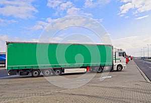 A row of trucks in a parking lot. In the foreground a truck with a semi-trailer. Green tarpaulin