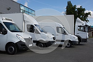 Row of white delivery and service van, trucks and cars in front of a factory and warehouse
