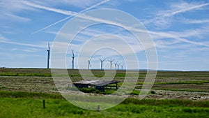 Row of wind turbines at a distance spinning in lush green field under blue cloudy sky