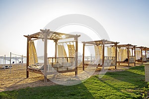 Row of wooden furniture for rest with curtains, deck chair bed, gazebo on sand beach with grass lawn, tropical sea luxury resort.