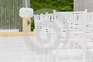 Rows of empty white chairs sitting on a wooden floor. Wedding chairs with flowers at ceremony outdoors