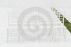 Rows of empty white chairs sitting on a wooden floor. Wedding chairs with flowers at ceremony outdoors