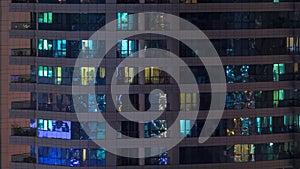 Rows of glowing windows with people in apartment building at night.