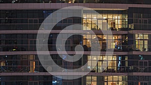 Rows of glowing windows with people in apartment building at night.