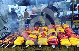 Rows of red, yellow, and orange bell peppers on skewers being grilled outdoors at a food market