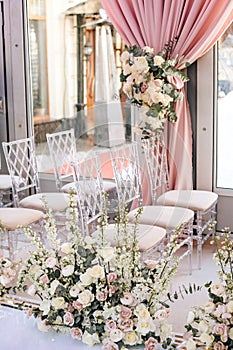 Rows of transparent chairs decorated with flower compositions of roses and white buttercups in the wedding ceremony area
