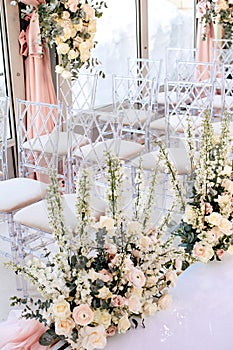 Rows of transparent chairs decorated with flower compositions of roses and white buttercups in the wedding ceremony area