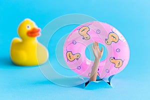 Rubber duck next to doll arm holding inflatable pool float and emerging from blue paper background. Drowning minimal creative abst