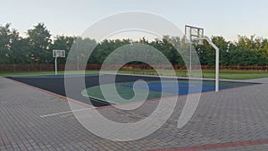The rubberized basketball court is located among grass lawns and concrete paved areas, next to trees and surrounded by a metal fen