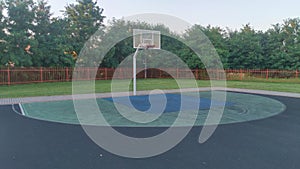 The rubberized basketball court is located among grass lawns and concrete paved areas, next to trees and surrounded by a metal fen