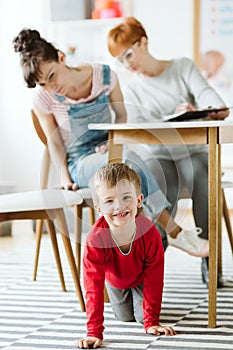 Rude kid sitting under the table during therapy for ADHD with his mother and professional therapist