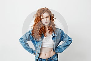 Rude young girl with red curly hair looking at camera brutally with arms akimbo over white background.