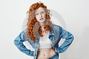 Rude young girl with red curly hair looking at camera brutally with arms akimbo over white background.