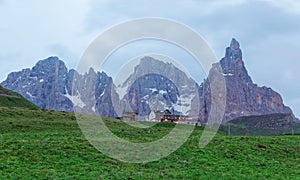 Rugged Cimon della Pala among peaks of Pale di San Martino mountains with a hotel on green grassy hill in Passo Rolle