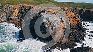 Rugged coastal cliff washing by stormy foamy ocean sunny day. Drone rough sea